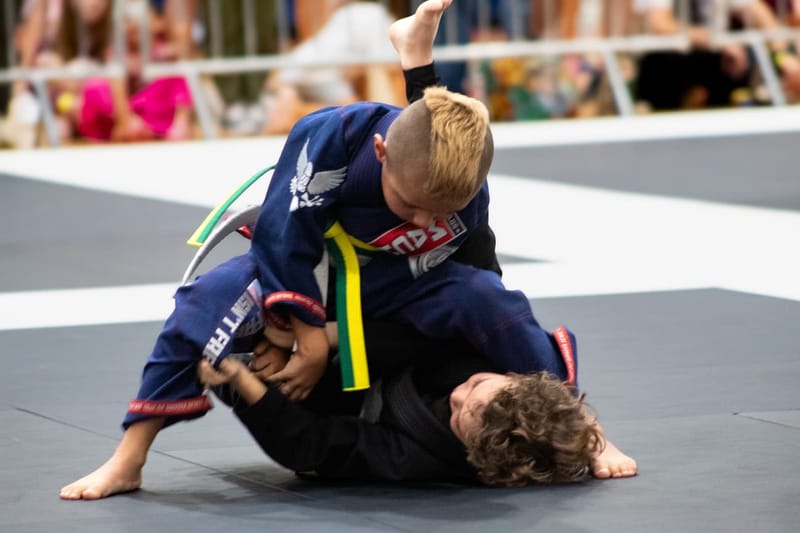 A blond boy in a navy gi controls his opponent from the top during a jiu-jitsu match.