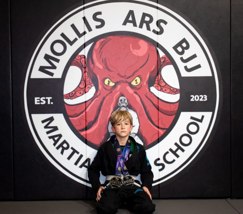 A blond boy in a black gi sits holding two grappling medals in front of the Mollis Ars BJJ logo wall.