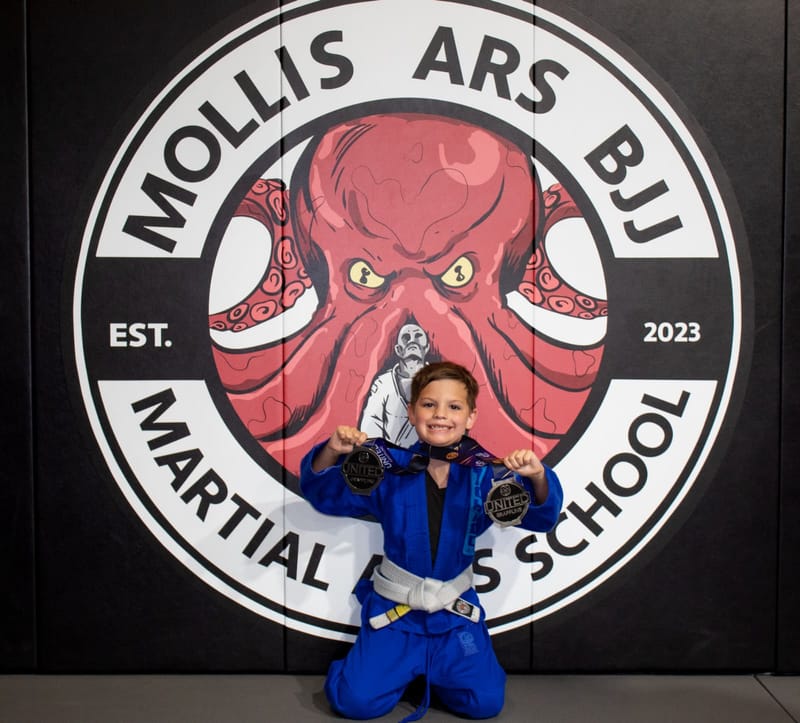 A grinning boy in a blue gi kneels holding up two medals in front of the octopus logo wall.