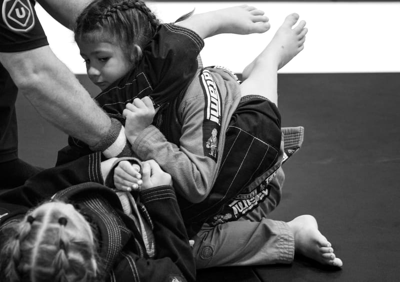 A young child controls a training partner from the mount in a black-and-white gi jiu-jitsu photo.