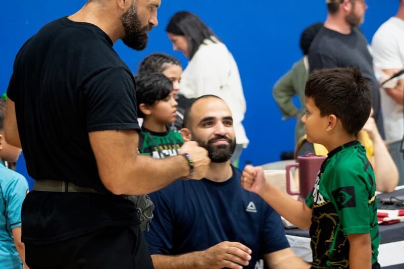 A coach fist-bumps a smiling young athlete in a green rash guard at a tournament.