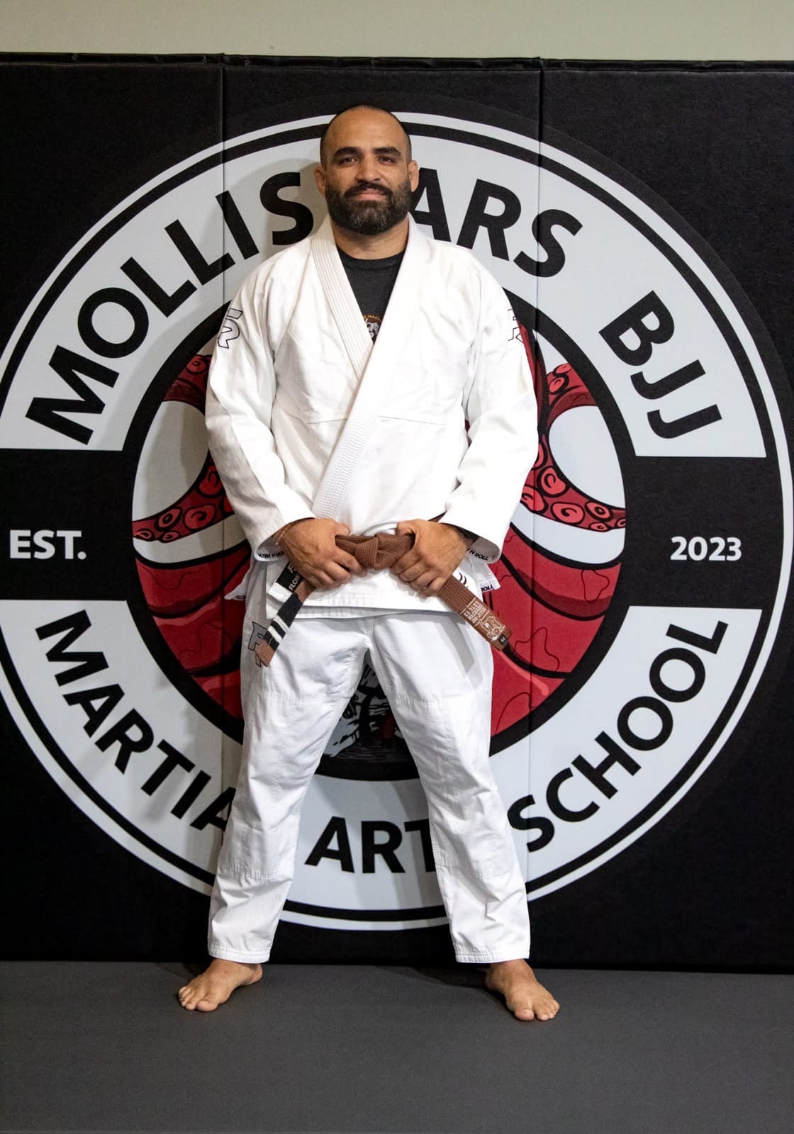 Dhavid Antunez, founder and head instructor at Mollis Ars BJJ, in his gi holding his brown belt in front of the academy logo wall