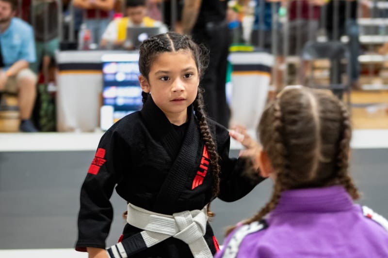 A focused young girl in a black gi squares off against an opponent at the start of a match.