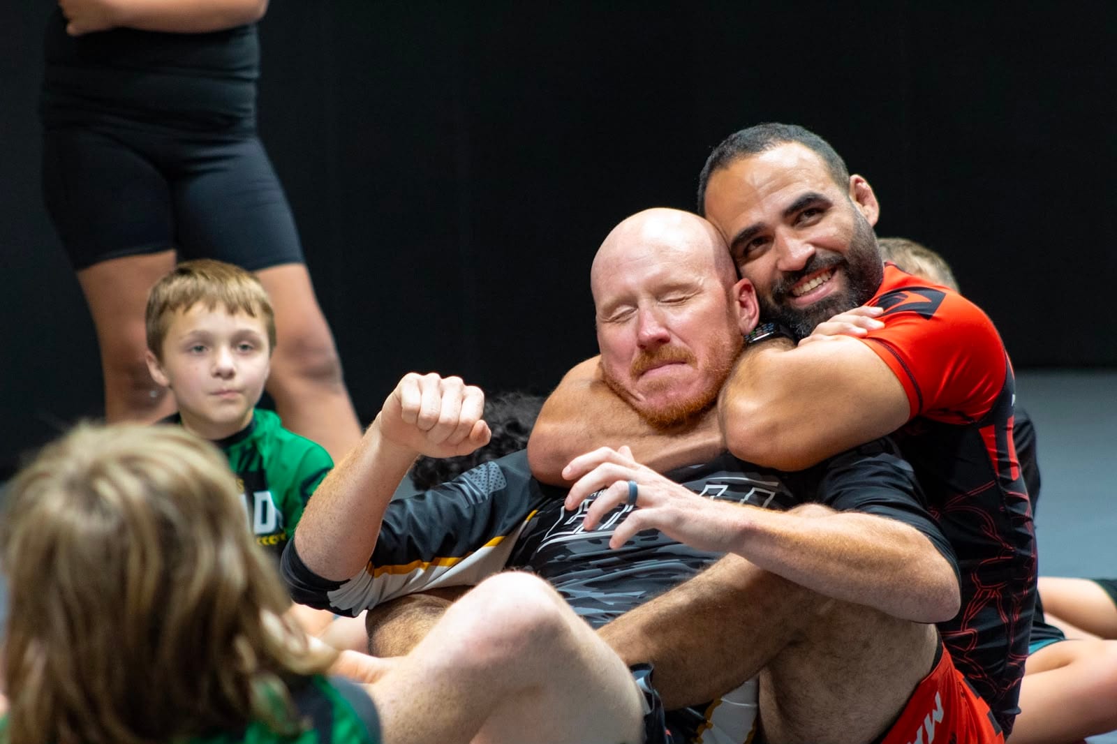 Two Mollis Ars BJJ training partners embracing after a roll while kids look on