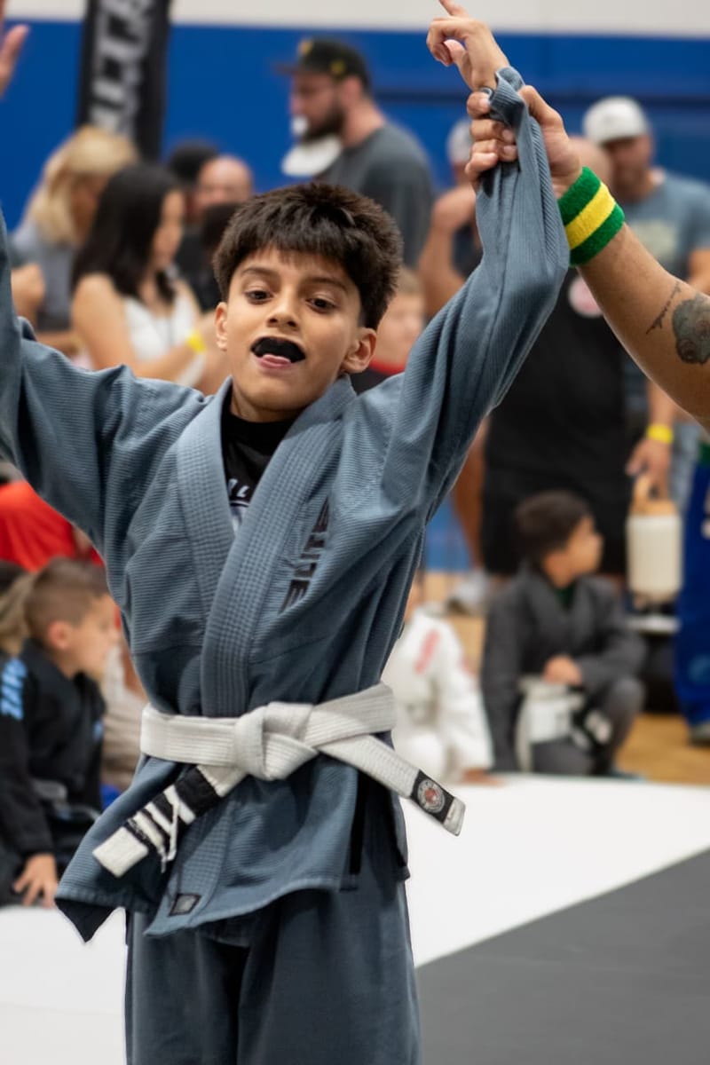 A young boy in a gray gi and white belt has his arm raised in victory by a referee at a jiu-jitsu tournament.