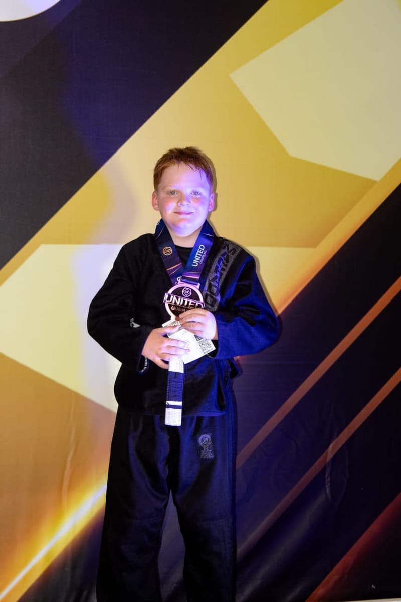A smiling red-haired boy in a black gi holds his medal in front of a tournament backdrop.