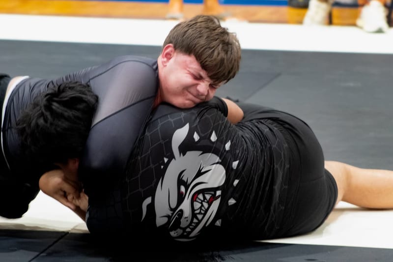 Two grapplers in black no-gi gear scramble on the mat as one applies pressure from the top.