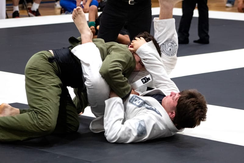 An older teen in a green gi drives to pass the guard of an opponent in a white gi during competition.