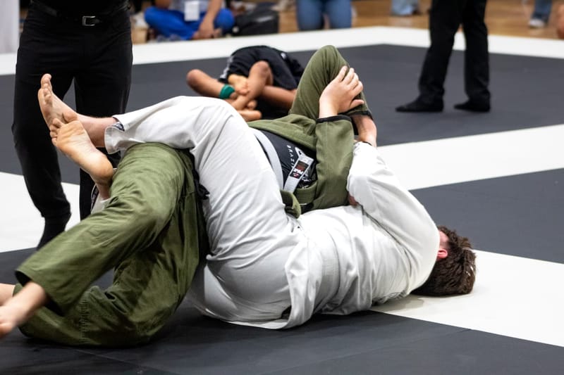 Two adult men grapple on the mat as one in a green gi works to pass the other’s guard.