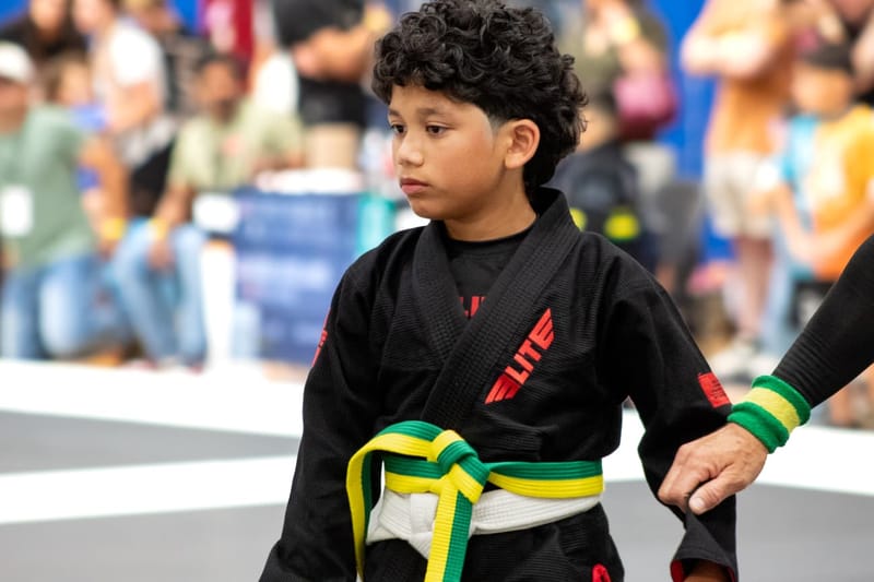 A young boy in a black gi with a green belt waits at the edge of the mat beside his coach.