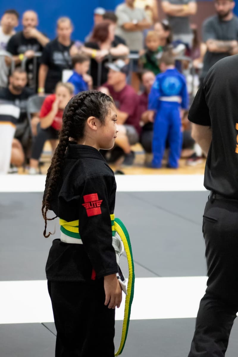 A young girl in a black gi with braided hair stands mat-side alongside her coach before a match.
