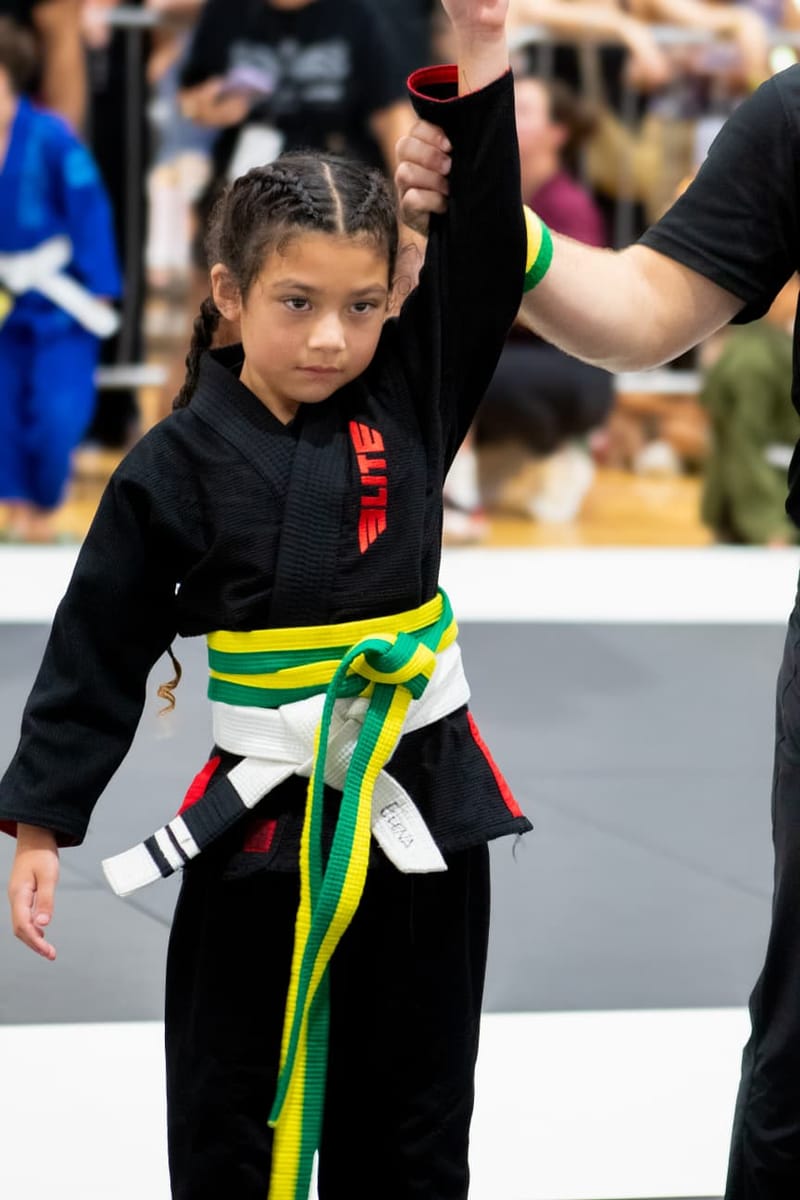 A young girl in a black gi has her hand raised in victory after winning her jiu-jitsu match.