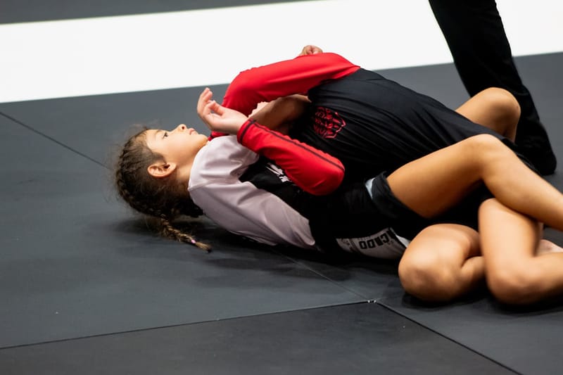 A young girl in a red rashguard works from her back during a no-gi grappling match.