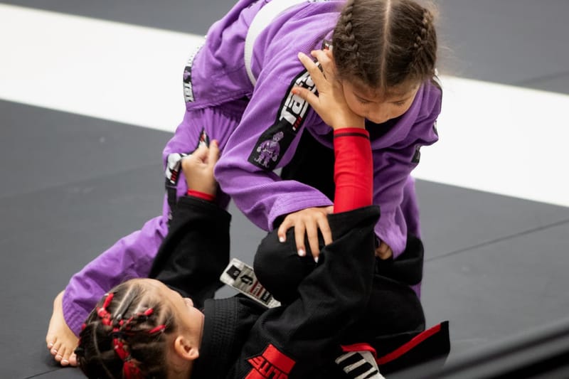 A young girl in a purple gi works to pass the guard of another girl in a black gi during a kids match.