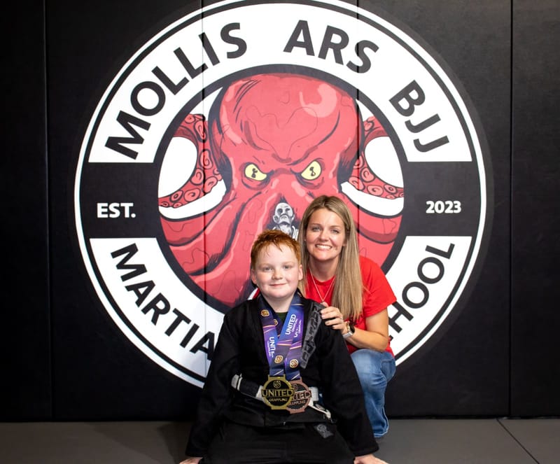 A red-haired boy in a black gi wearing grappling medals poses with his smiling mother at the logo wall.